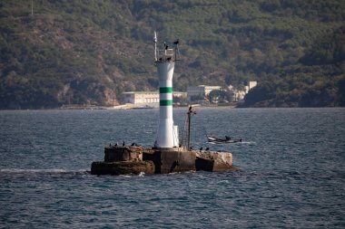 İstanbul, Türkiye - Boğaz 'ın kayalık bir adasında, arka planda ağaçlık bir kıyı şeridi ve küçük bir teknenin geçtiği yeşil çizgili küçük bir seyir feneri bulunuyor.