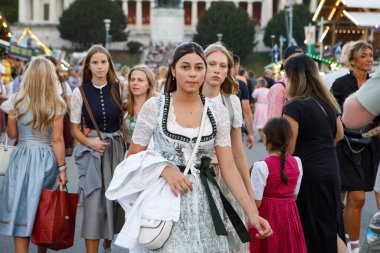Münih, Almanya - 21 Eylül 2023: Bir grup zarif geleneksel Bavyera Dirndls giysisi giymiş genç kadın, Bavyera 'daki festival alanlarında yürüyor, Bavyera heykeli ve Ruhmeshalle arka planda Oktoberfest sırasında görülüyor.