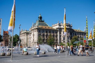Münih, Almanya - 21 Eylül 2023: Yayalar güneşli bir günde Stachus Meydanı 'ndaki tarihi neo-barok Adalet Sarayı önünde bir çeşmenin önünden geçtiler.