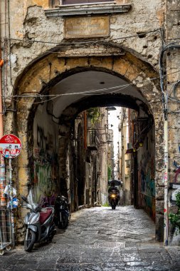 Napoli, İtalya - 4 Kasım 2022: Bir motosiklet Pendino ve San Lorenzo bölgeleri arasındaki dar tarihi bir sokakta modern duvar yazısıyla kaplı bir taş kemer yolundan geçiyor..
