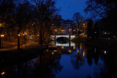 Riga, Letonya 'da, sonbahar parkı ağaçları ve parlayan sokak lambalarıyla çevrili mavi saat alacakaranlığı sırasında Pilsetas Kanallarının sakin sularına tarihi bir taş köprü yansıyor.