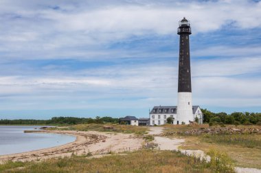 İkonik siyah-beyaz Sorve deniz feneri Estonya 'nın Saaremaa kentindeki bulutlu mavi gökyüzünün altında Baltık Denizi kıyısındaki çimenli bir çayır üzerinde dimdik duruyor.