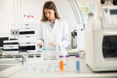Laboratuvarda koruyucu yüz maskesi takan genç bir bayan doktor elinde sıvı numunesi olan bir şişe tutuyor.