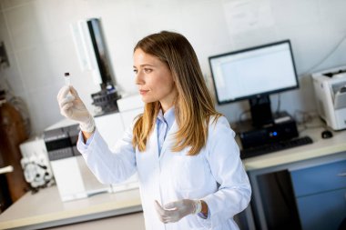 Young female scientist in a white lab coat preparing vial with a sample for an analysis on a gas chromatograph in biomedical lab clipart