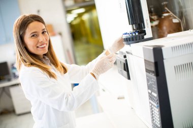 Beyaz önlüklü genç kadın bilim adamı biyomedikal laboratuardaki bir gaz kromatografisinin analizi için bir numuneyle birlikte şişeyi koyuyor.