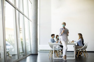 Koruyucu maskeli olgun bir iş kadını ekibinin önünde dijital tablet kullanıyor.