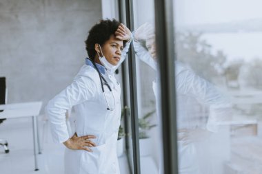 Siyahi kadın doktor, sağlık ofisinin penceresinin önünde negatif bir ifadeyle dikiliyor.