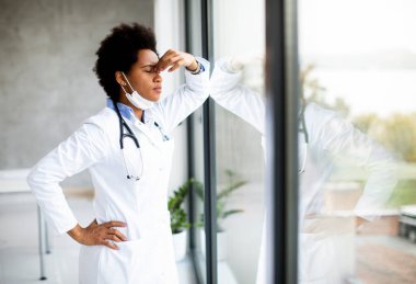 Siyahi kadın doktor, sağlık ofisinin penceresinin önünde negatif bir ifadeyle dikiliyor.