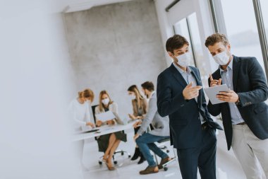 Yüz koruyucu maskeli iki yakışıklı genç iş adamı ofiste kağıt planlarla tartışıyorlar.