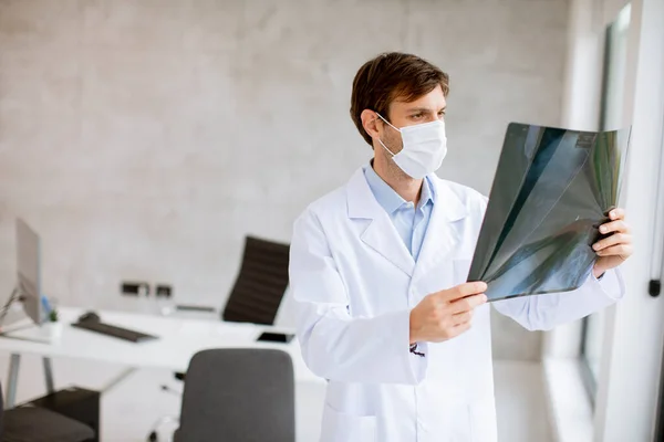 Νεαρός Γιατρός Προστατευτική Μάσκα Προσώπου Που Εξετάζει Ακτινογραφία Στο Γραφείο — Φωτογραφία Αρχείου