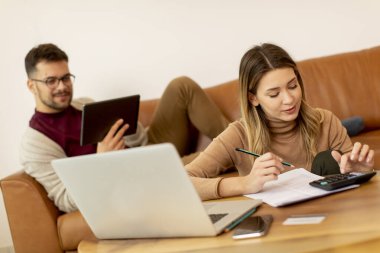 Genç bir kadın dizüstü bilgisayar kullanıyor ve genç bir adam evdeki kanepede otururken dijital tablet kullanıyor.