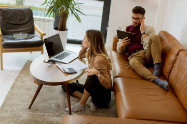 Genç bir kadın dizüstü bilgisayar kullanıyor ve genç bir adam evdeki kanepede otururken dijital tablet kullanıyor.