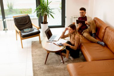 Genç bir kadın dizüstü bilgisayar kullanıyor ve genç bir adam evdeki kanepede otururken dijital tablet kullanıyor.