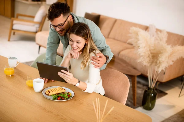 デジタルタブレットを使用して素敵な若いカップルとキッチンで朝食を持っています — ストック写真