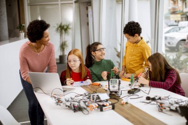 Bir grup mutlu çocuk ve dizüstü bilgisayarı olan Afrika kökenli Amerikalı kadın fen öğretmenleri robot sınıfında elektrikli oyuncaklar ve robotlar programlıyorlar.