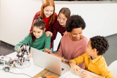 Bir grup mutlu çocuk ve dizüstü bilgisayarı olan Afrika kökenli Amerikalı kadın fen öğretmenleri robot sınıfında elektrikli oyuncaklar ve robotlar programlıyorlar.