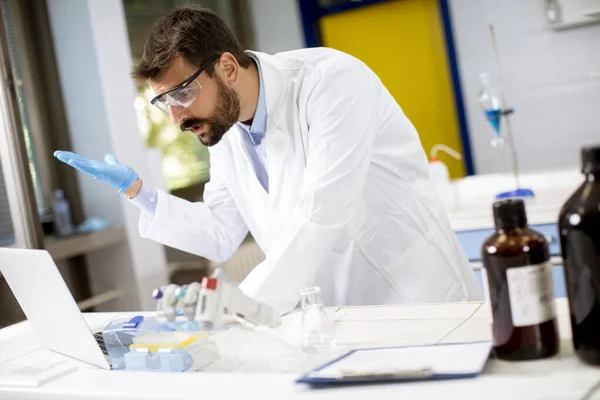 年轻英俊的研究人员穿着白色的实验室外套 坐在实验室里用笔记本电脑工作 — 图库照片
