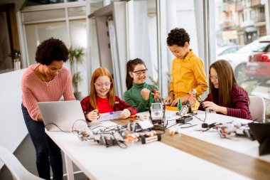 Bir grup mutlu çocuk ve dizüstü bilgisayarı olan Afrika kökenli Amerikalı kadın fen öğretmenleri robot sınıfında elektrikli oyuncaklar ve robotlar programlıyorlar.