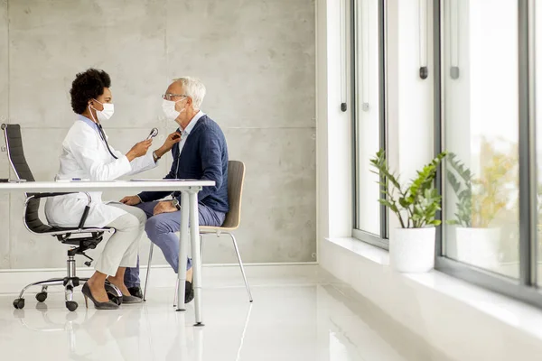 Senior man with protective facial mask receive news from black female doctor in the office