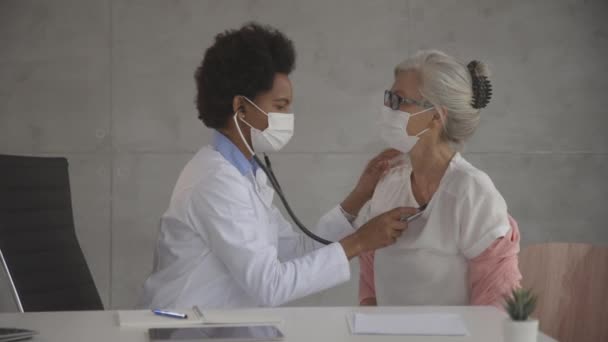 有护肤面罩的老年妇女在办公室接受黑人女医生的体格检查 — 图库视频影像