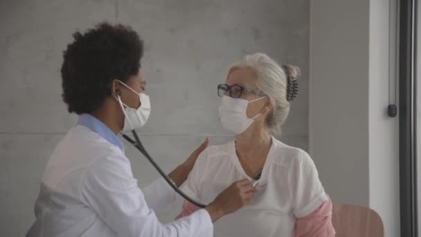 Mulher sênior com máscara facial protetora tendo um exame médico por médico feminino preto no escritório — Vídeo de Stock