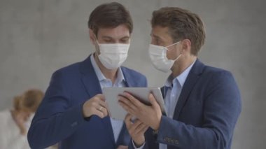 Yüz koruyucu maskeli iki yakışıklı iş adamı ofiste dijital tabletle tartışıyor.