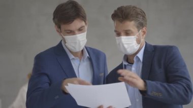 Yüz koruyucu maskeli iki yakışıklı genç iş adamı ofiste kağıt planlarla tartışıyorlar.