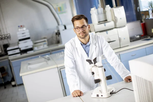 材料科学研究所の両眼顕微鏡を使った白衣を着たハンサムな若い科学者 — ストック写真
