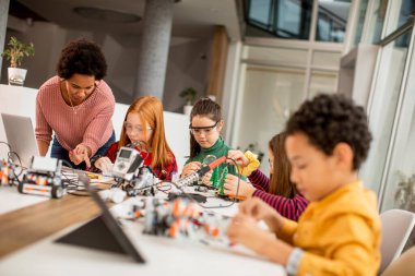 Bir grup mutlu çocuk ve dizüstü bilgisayarı olan Afrika kökenli Amerikalı kadın fen öğretmenleri robot sınıfında elektrikli oyuncaklar ve robotlar programlıyorlar.
