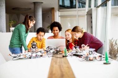 Bir grup mutlu çocuk ve dizüstü bilgisayarı olan Afrika kökenli Amerikalı kadın fen öğretmenleri robot sınıfında elektrikli oyuncaklar ve robotlar programlıyorlar.