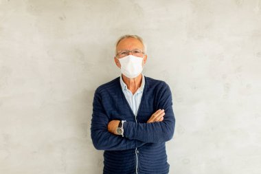 Kıdemli adam yüz koruyucu maske takıyor ve kollarını kavuşturarak duvarın yanında duruyor.