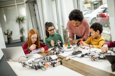 Bir grup mutlu çocuk ve dizüstü bilgisayarı olan Afrika kökenli Amerikalı kadın fen öğretmenleri robot sınıfında elektrikli oyuncaklar ve robotlar programlıyorlar.