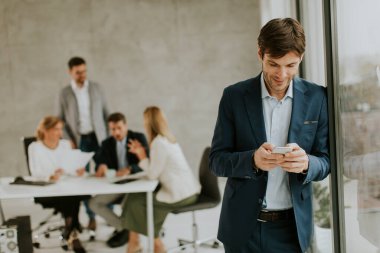 Yakışıklı genç iş adamı ofisin önünde ekibinin önünde kendinden emin duruyor ve cep telefonu kullanıyor.
