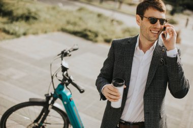 Bisikletin yanında kahve fincanıyla cep telefonu kullanan yakışıklı bir iş adamı.