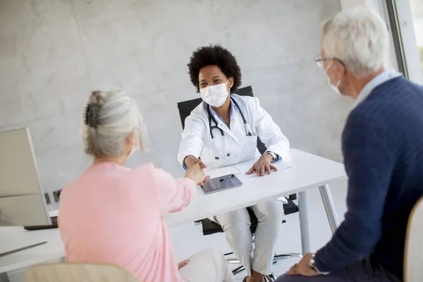 Senior couple with protective facial masks receive news from black female doctor in the office