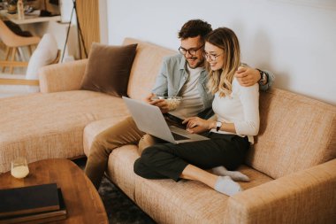 Yakışıklı genç çift evdeki kanepede otururken birlikte dizüstü bilgisayar kullanıyorlar.
