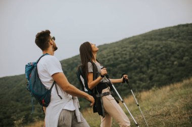 Bir yaz gününde yeşil tepelerde sırt çantalarıyla yürüyen gülümseyen genç bir çift.
