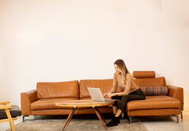 Genç ve güzel bir kadın evdeki kanepede dizüstü bilgisayar kullanıyor.