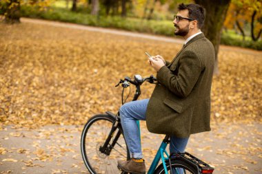 Güz parkında elektrikli bisikletle cep telefonu kullanan yakışıklı genç adam.
