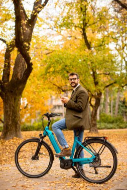 Güz parkında elektrikli bisikletle cep telefonu kullanan yakışıklı genç adam.