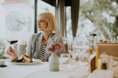 A woman sits at a stylish dining table, savoring a glass of red wine. She is dressed in a light gray blazer and striped top, with a gourmet meal and flowers on the table.
