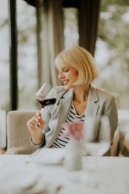 A smiling woman in a light blazer examines her glass of red wine at a stylish restaurant. Natural light fills the space, enhancing the inviting atmosphere.