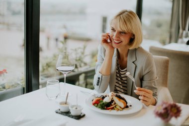 A woman sits at a table, savoring her delicious meal with a glass of red wine. She smiles contentedly while enjoying the upscale ambiance of the restaurant with large windows.