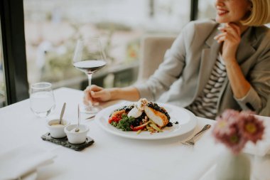 A woman enjoys a gourmet meal of salmon with fresh vegetables while sipping red wine. She smiles, sitting in an elegant restaurant with natural light streaming in.