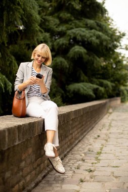 A blond woman with a blazer and white pants sits on a brick wall outside. She looks at her smartphone and smiles. Green trees are in the background.