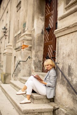 A woman sits on the steps of an old building, engrossed in her book. She wears light clothing and enjoys the warm afternoon, surrounded by historic architecture.