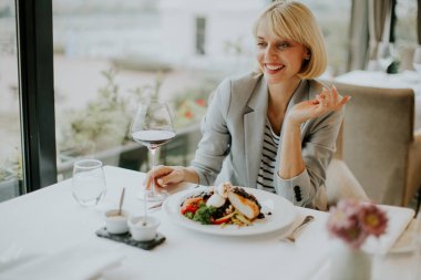 A woman sits at a table in a modern restaurant, savoring a beautifully presented gourmet dish accompanied by wine. She looks relaxed and happy.