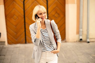 A smiling woman dressed in a striped shirt and blazer is walking on the sidewalk while talking on her phone. The background features a wooden door and light-colored walls.