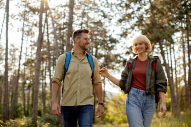 A man and woman walk side by side in a tranquil forest, sharing smiles while exploring nature together on a sunny day.
