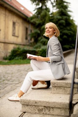 A young woman sits on stone steps, smiling as she reads a book. Dressed in a light gray blazer and white pants, she enjoys a moment in a peaceful, scenic location.